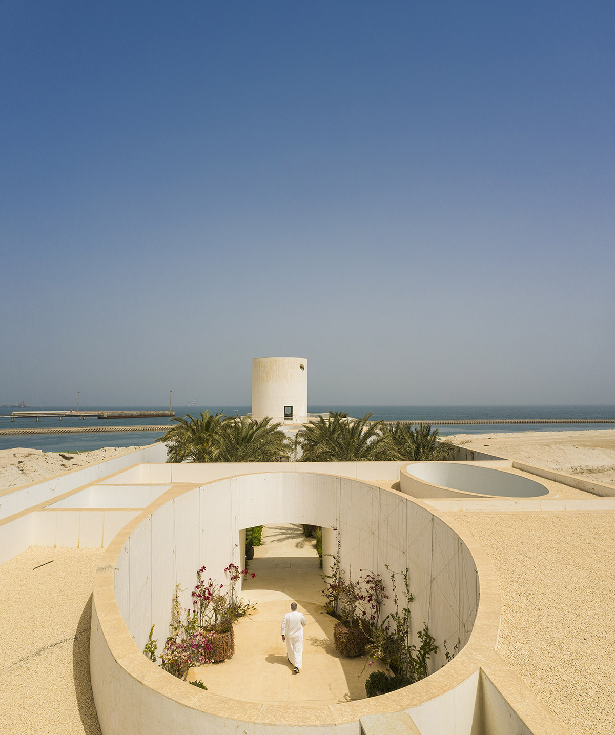 White Fortress by TAEP/AAP is a linear house that features a tall cylindrical lighthouse on a beach 