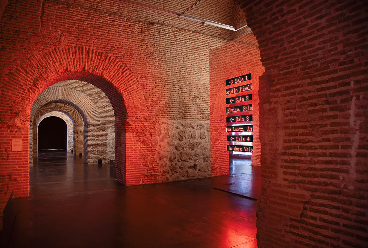 Exhibition design at Condeduque Center’s vaulted hall plays with red light, mirrors and reflections